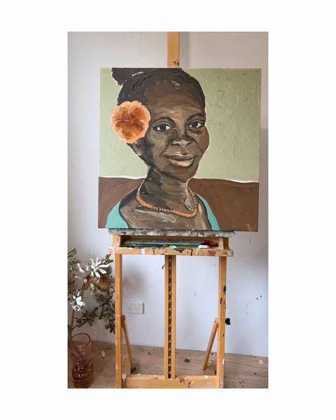 A painted portrait of a smiling woman with dark skin, a teal top, and a peach Hibiscus flower in her hair, displayed on a wooden artist easel in a studio 